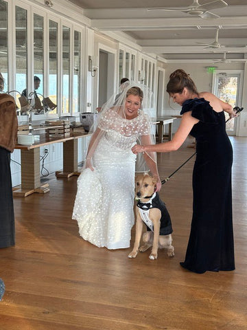 Dog wearing a floral collar standing beside a bride and groom at an outdoor wedding